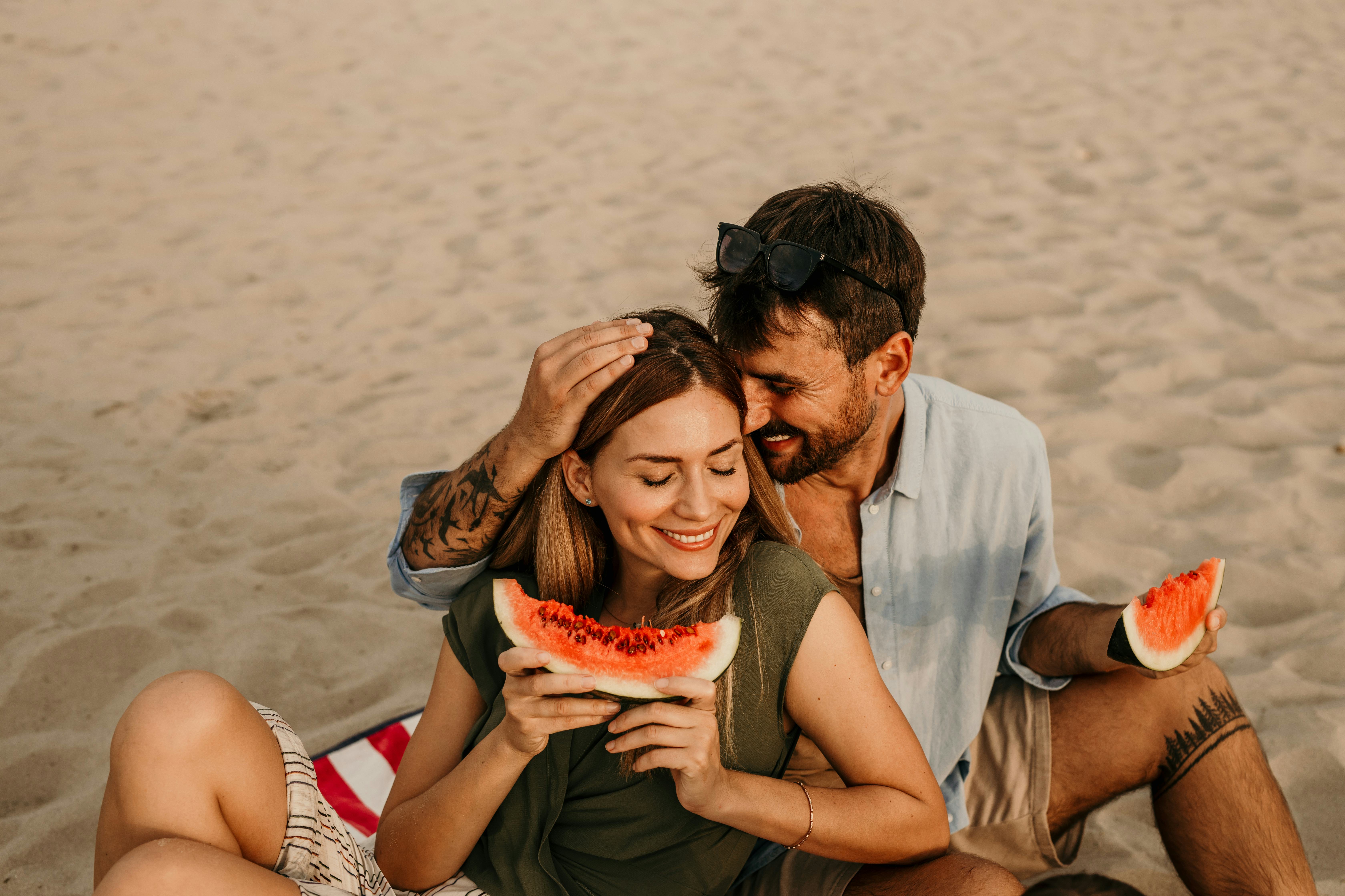 A couple sitting on the beach together eating watermelon and giving each other fun pet names.