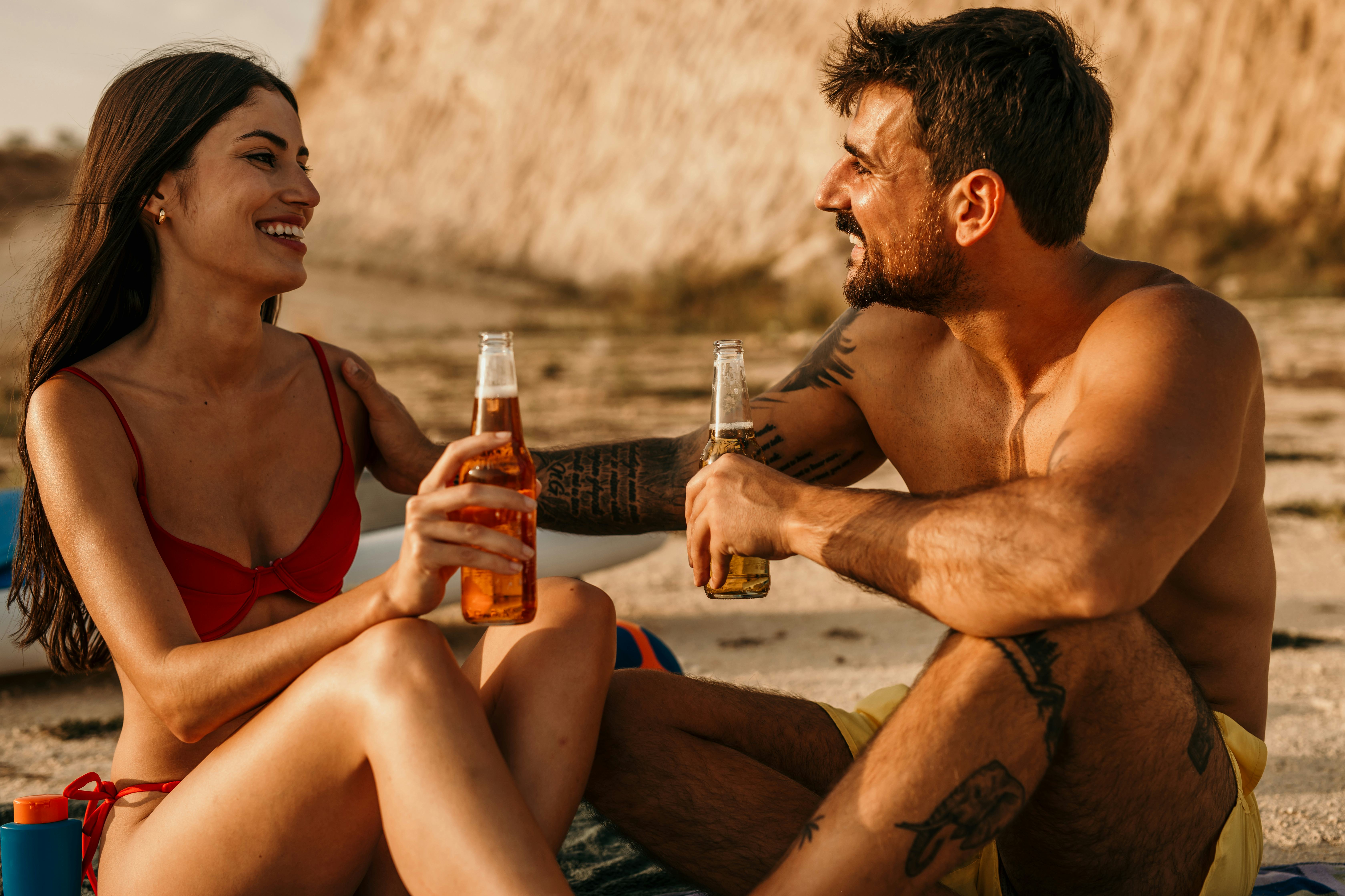 A girlfriend making her boyfriend smile at the beach with sweet nicknames as they drink beers.