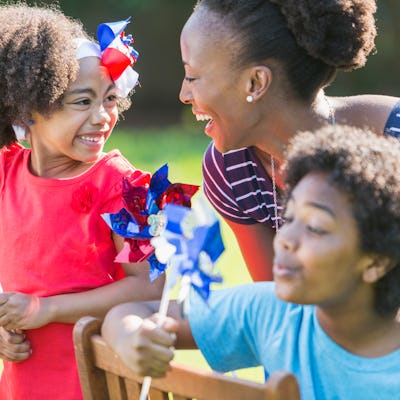 An African American mother with two mixed race children celebrating an American patriotic holiday, perhaps July 4th or Memorial Day. They are playing with red, white and blue pinwheels, smiling and laughing, outdoors on a bright, sunny day. The children are black, Asian and Hispanic. The focus is on the woman and girl who are looking at each other.