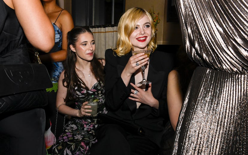 Two women at a social event; one in a floral dress holds a drink, the other in a black outfit laughs...