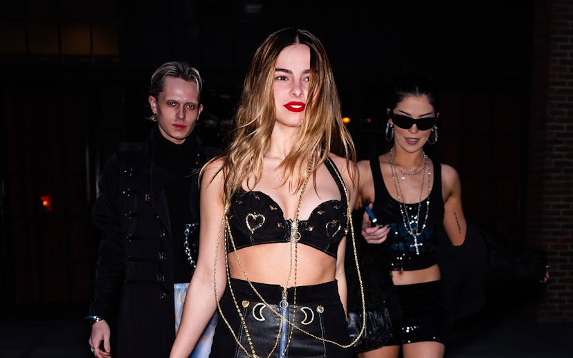 Three people walking on a city street at night, two women and one man, dressed in stylish, edgy outf...
