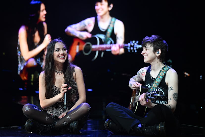 Two women on stage, one sitting and singing with a microphone, the other playing guitar, both smilin...