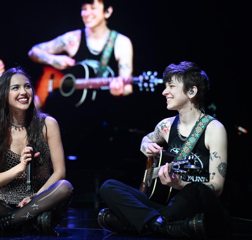 Two women on stage, one sitting and singing with a microphone, the other playing guitar, both smilin...