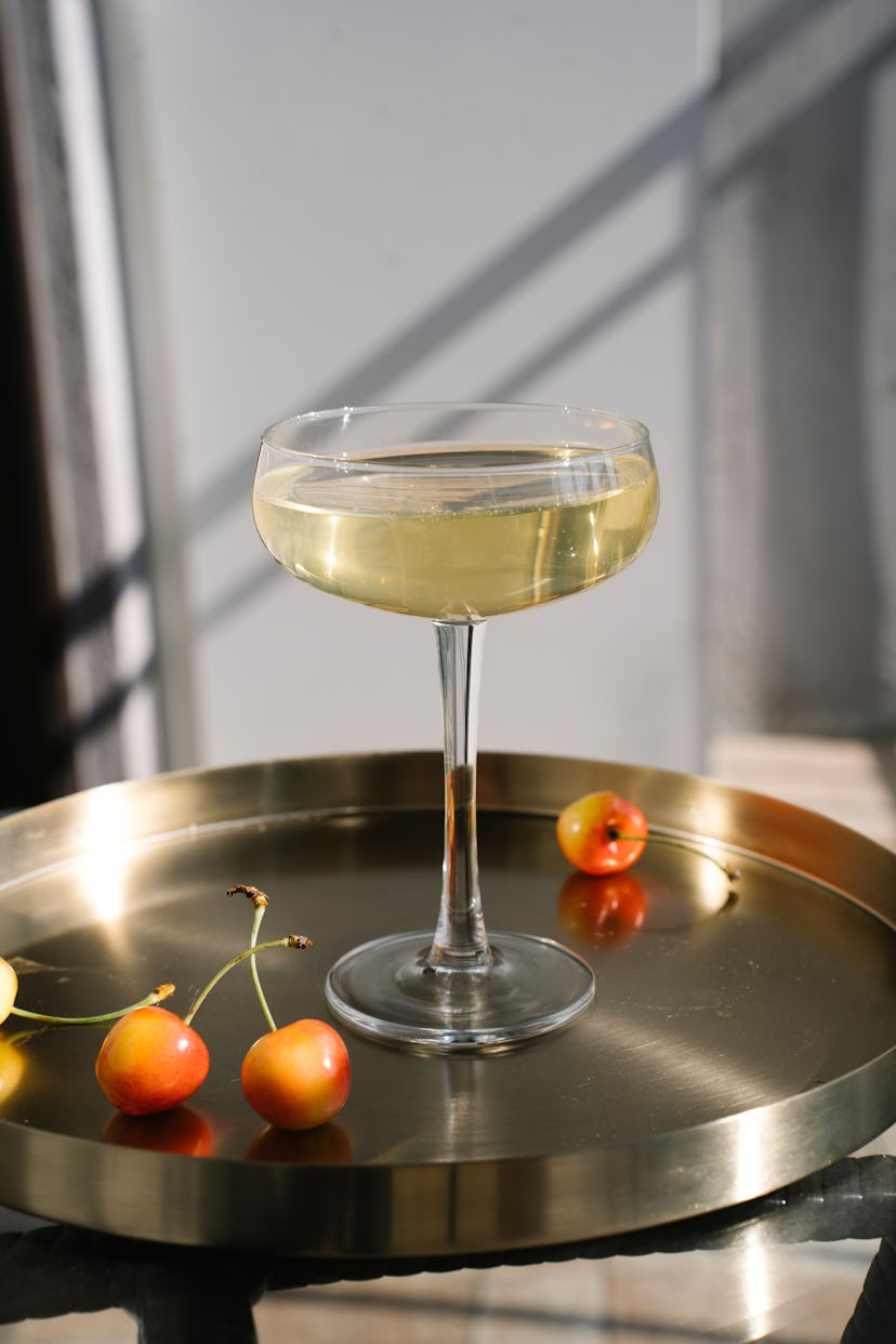 White wine in drinking glass and pink cherry fruits in golden tray on white table in bright sunlight