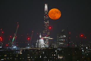 A large, orange moon rises behind the illuminated skyline of London, featuring St. Paul's and the Shard, at night.
