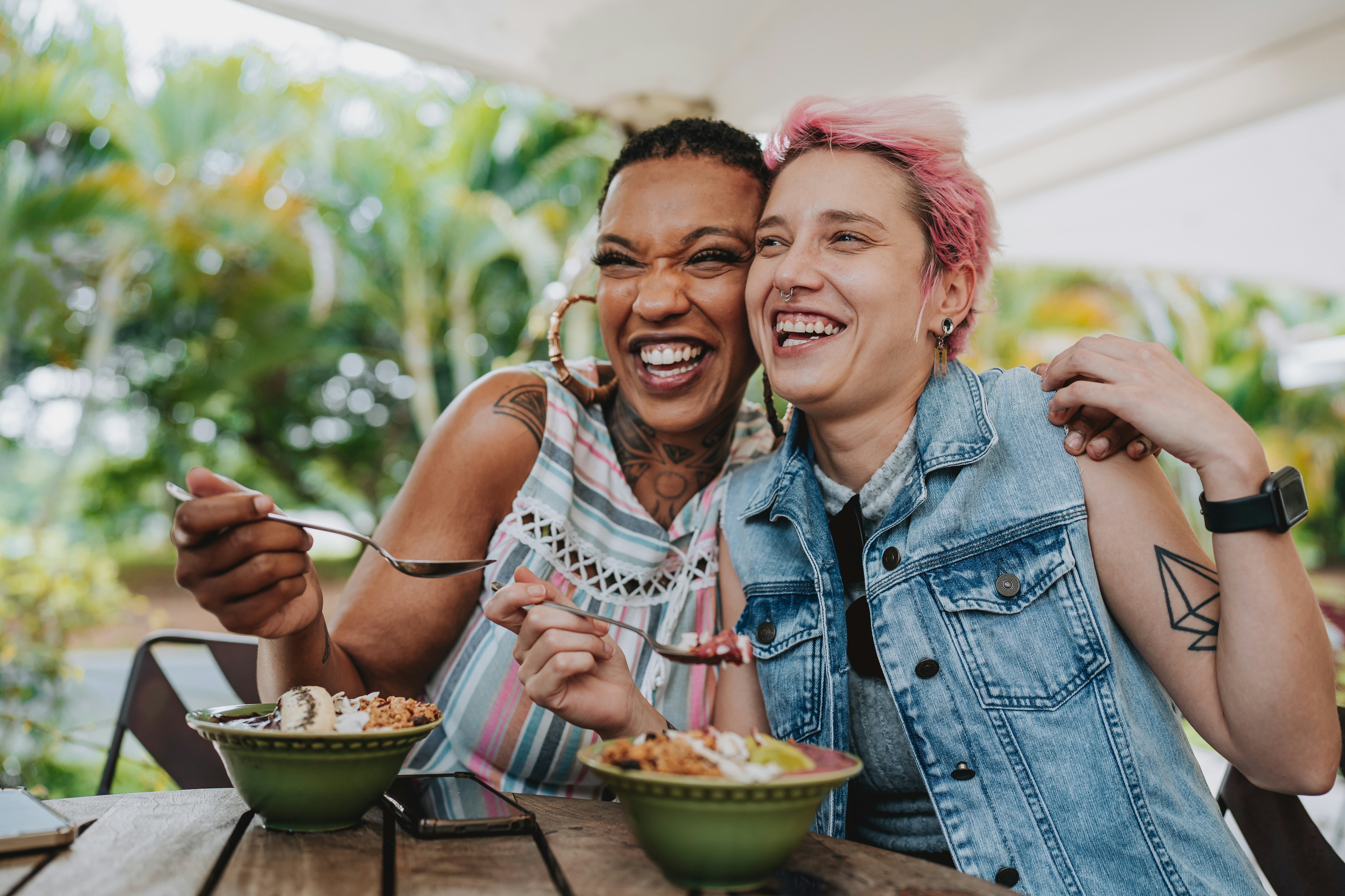 Two young female friends laughing at food riddles for adults and eating dessert in an outdoor restau&hellip;