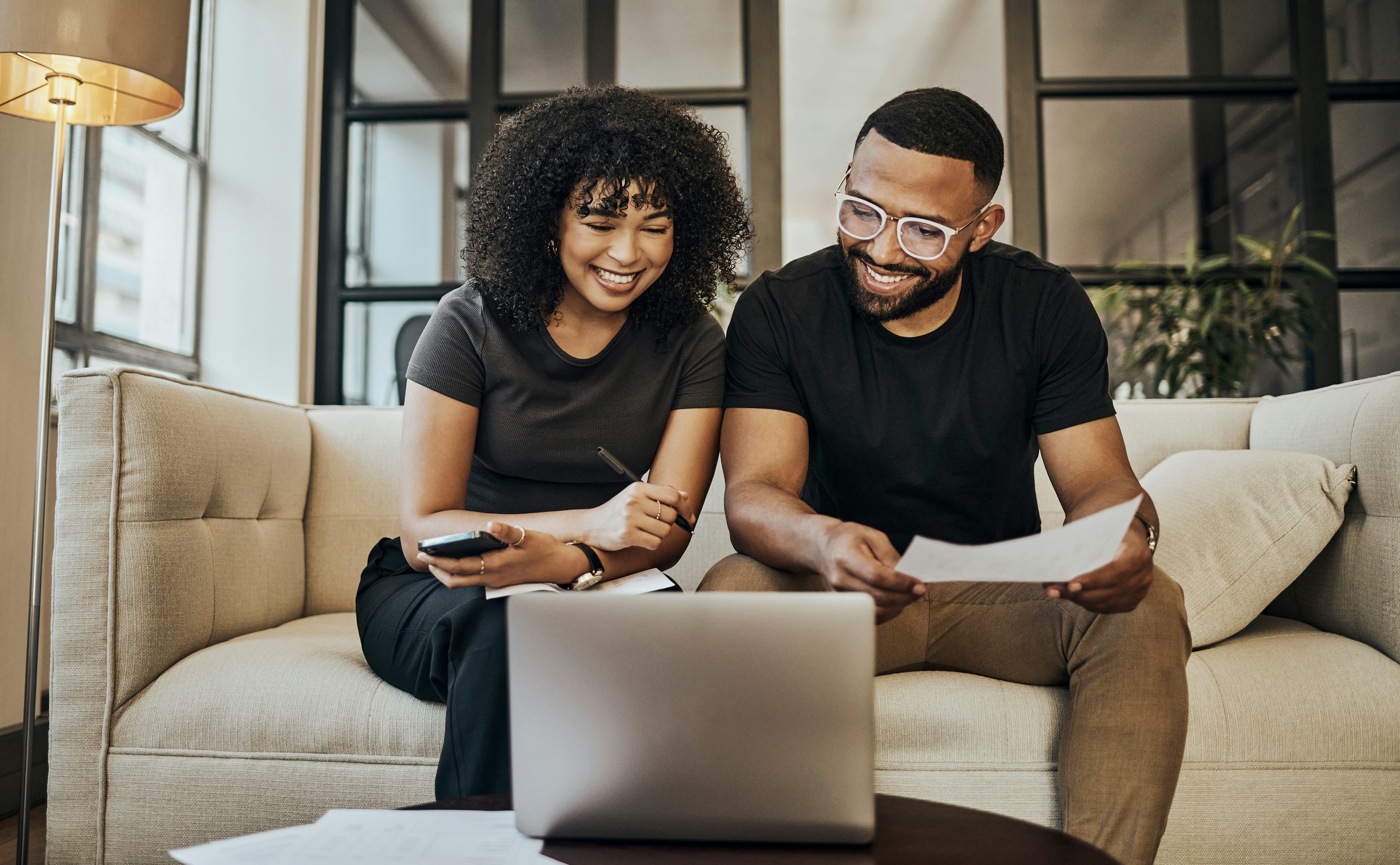 A young couple on their couch looking at their finances on their computer while telling riddles abou&hellip;