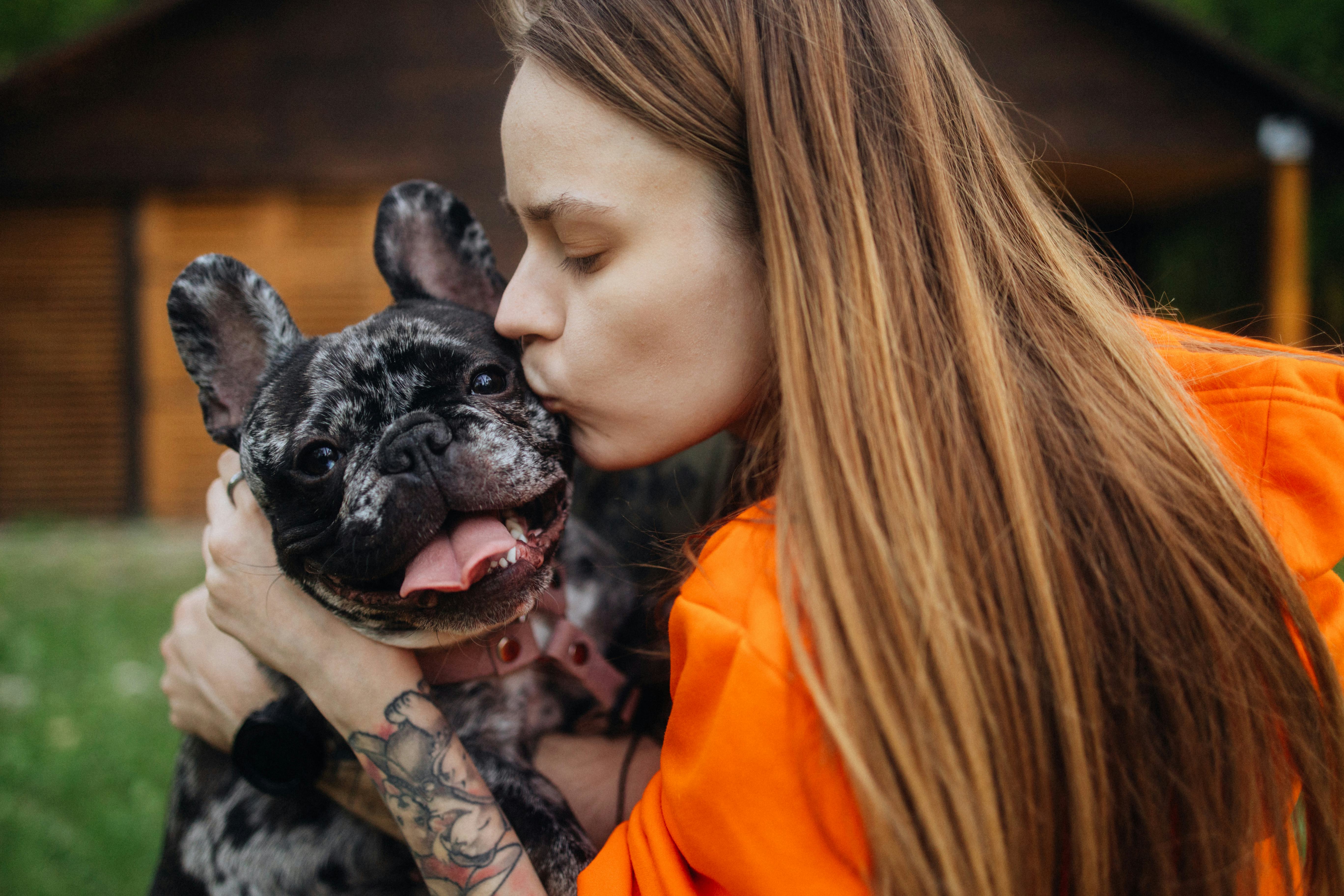 A young woman whispers silly adult animal riddles into her bulldog&rsquo;s ear and gives him kisses.