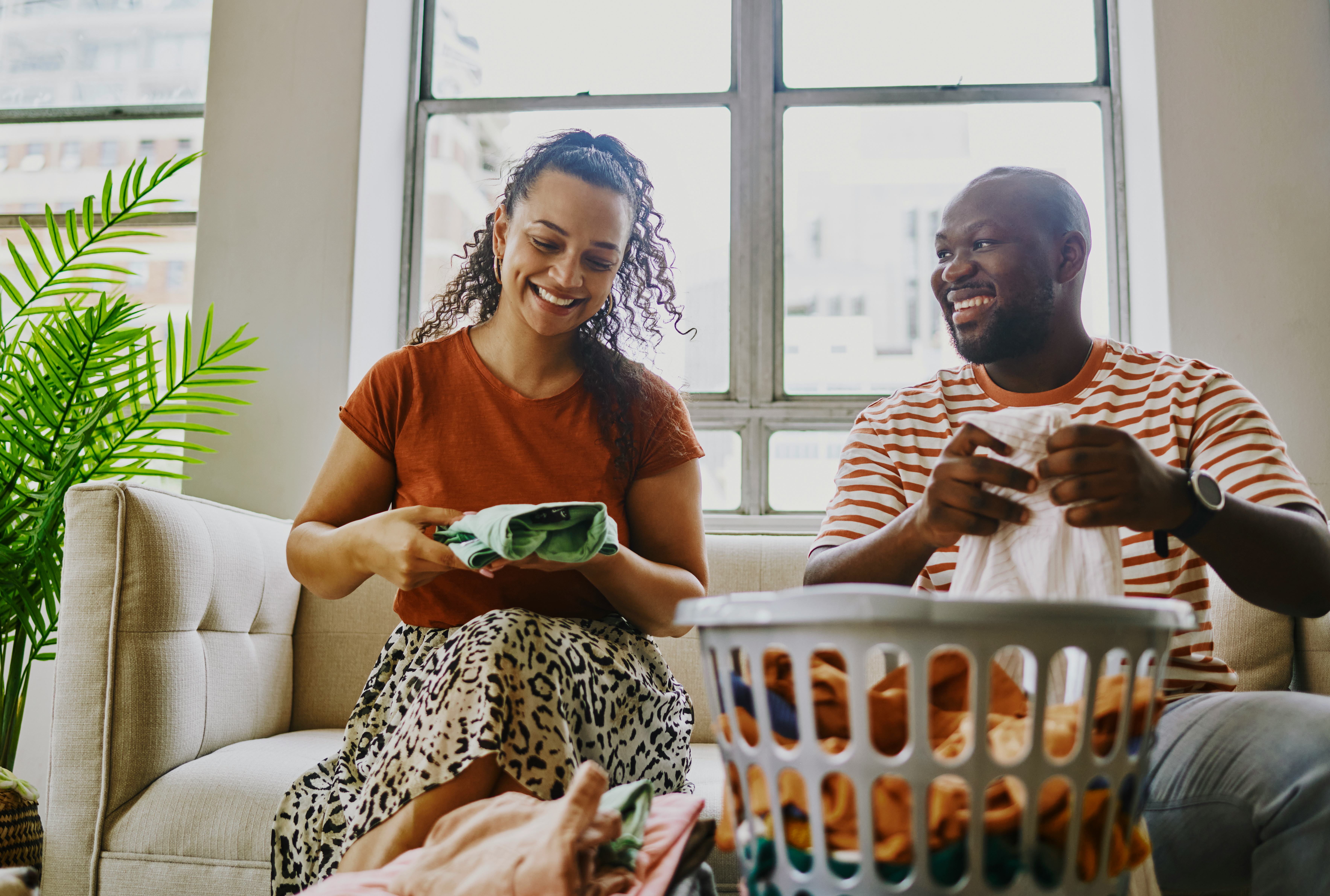 A young couple sits on their couch and tells each other riddles for adults while folding laundry.