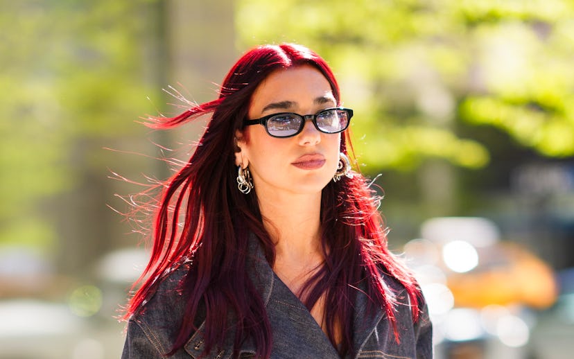 Woman with red hair and glasses wearing a denim jacket over a dress, holding a silver chain bag, wal...