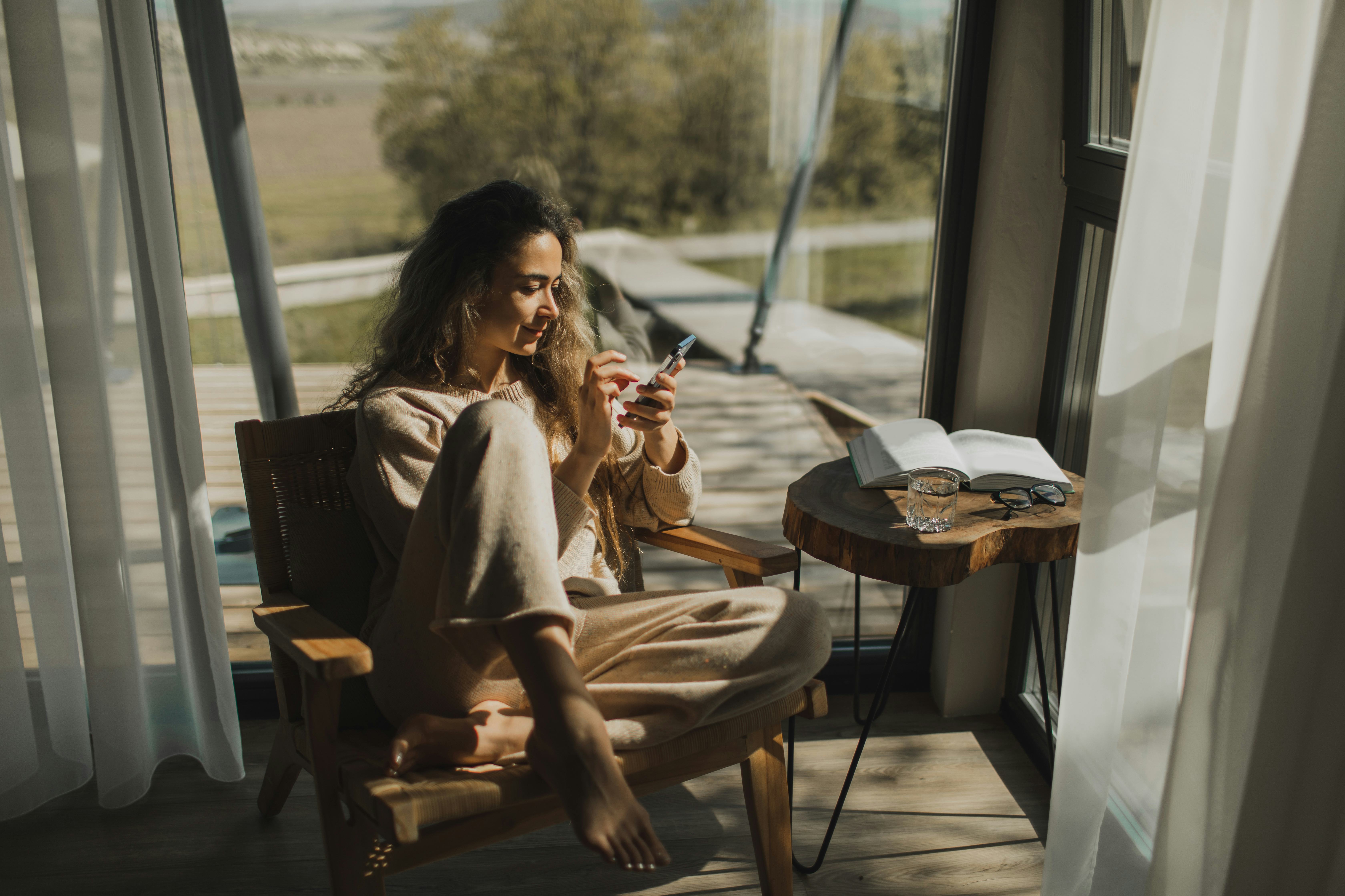 A young woman sits in a chair by a large window, looking for easy riddles for adults on her phone.