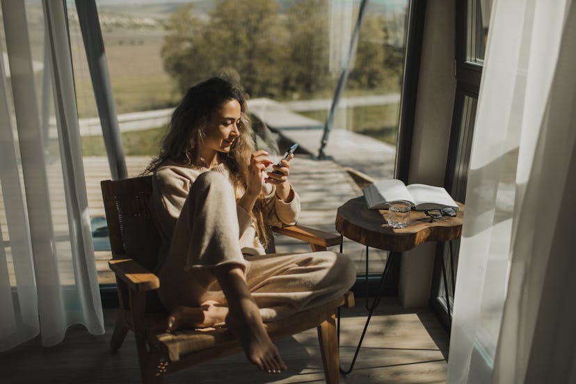 A young woman sits in a chair by a large window, looking for easy riddles for adults on her phone.