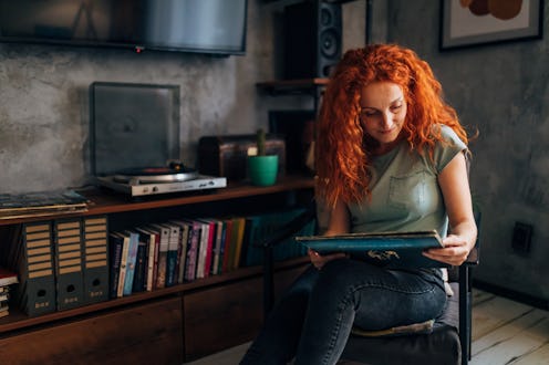 A woman sits in a chair in her living room and reads riddles for adults on her tablet in her lap.
