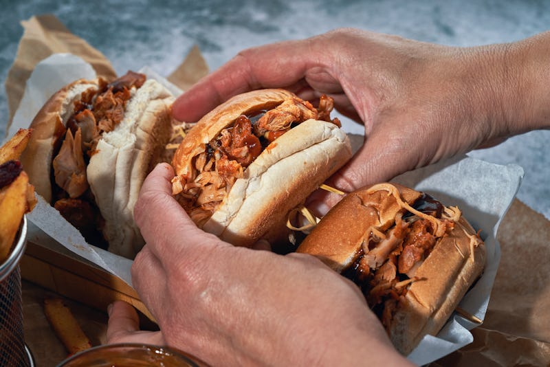 Hands holding three pulled pork sandwiches, with shredded meat heaping from soft buns, on a paper-li...