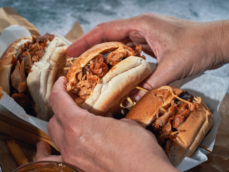 Hands holding three pulled pork sandwiches, with shredded meat heaping from soft buns, on a paper-li...