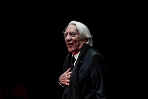 Elderly man in a black suit, standing on stage with a surprised expression, lit against a dark backg...