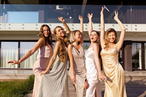 Portrait of excited young women wearing elegant dresses standing in front of villa and raising hands...