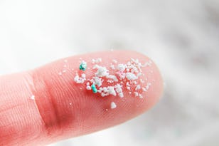 Close-up of a fingertip with white and green microplastic particles scattered on it, against a snowy background.