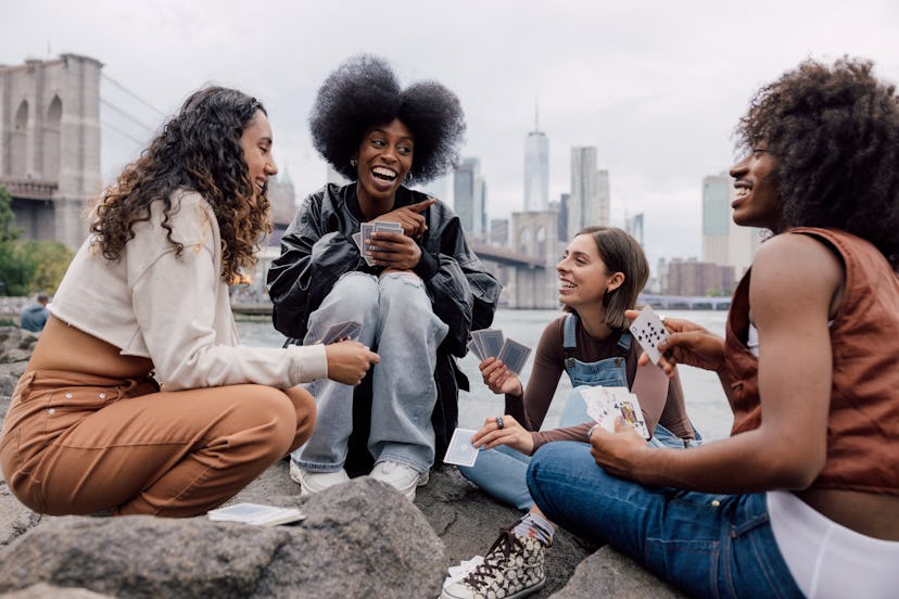 A group of four friends sitting down in front of the New York skyline playing cards and telling fun …