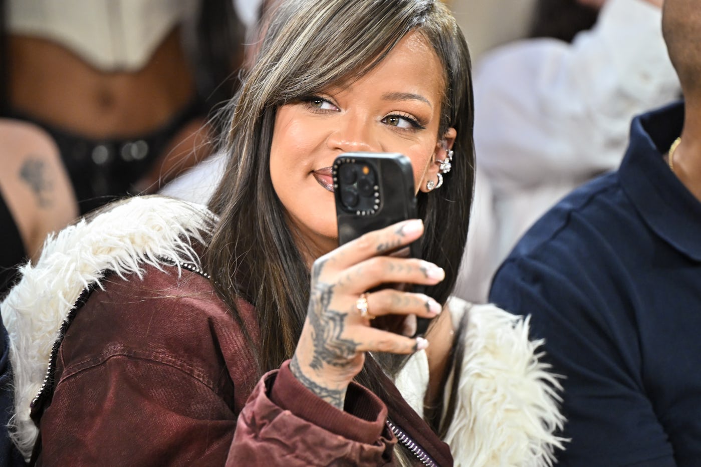 A woman in a white and burgundy jacket using a smartphone to take a photo, smiling with detailed tattoos on her hand.