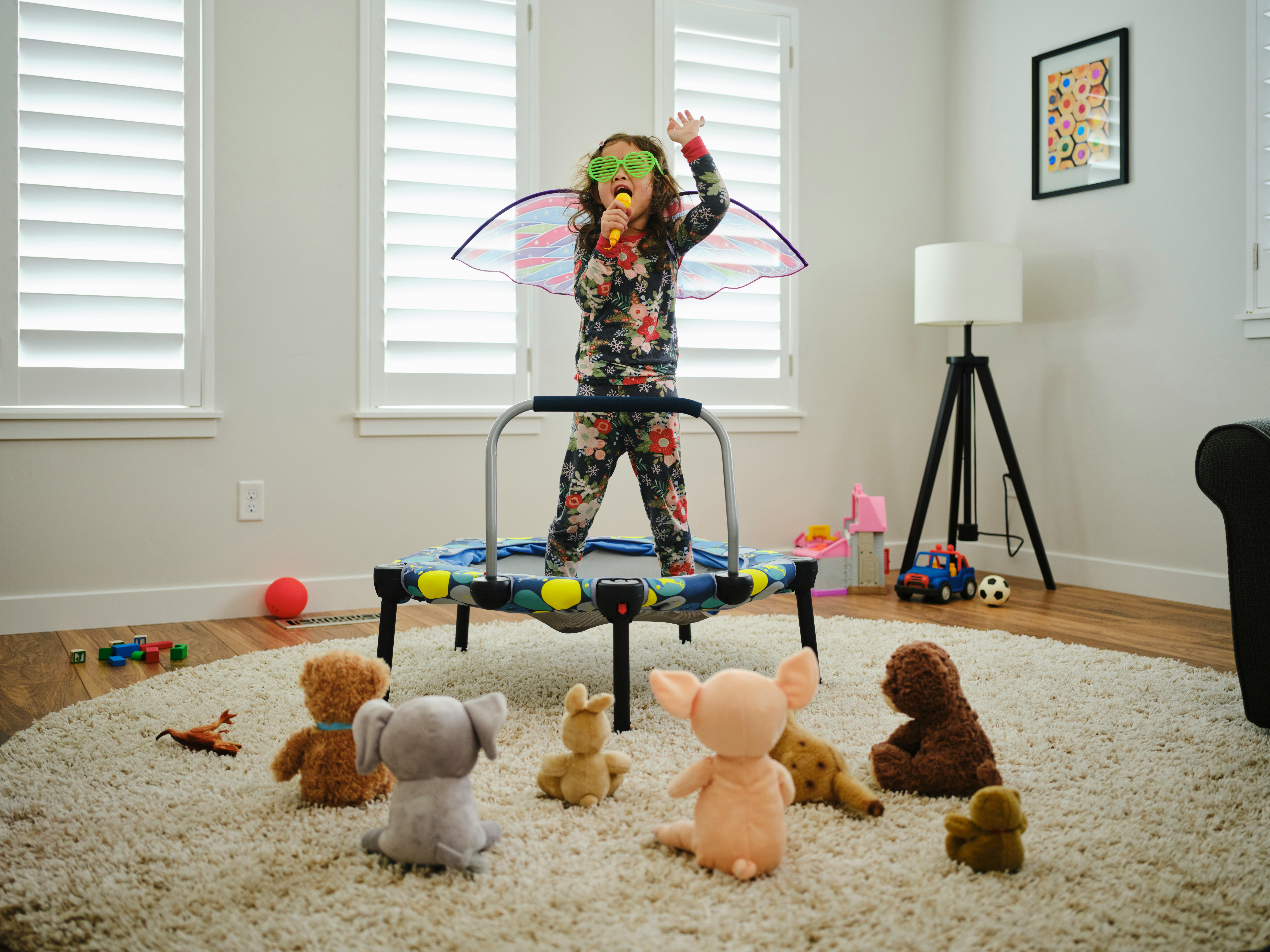 A toddler in butterfly wings putting on a concert for her stuffed animals in her living room.