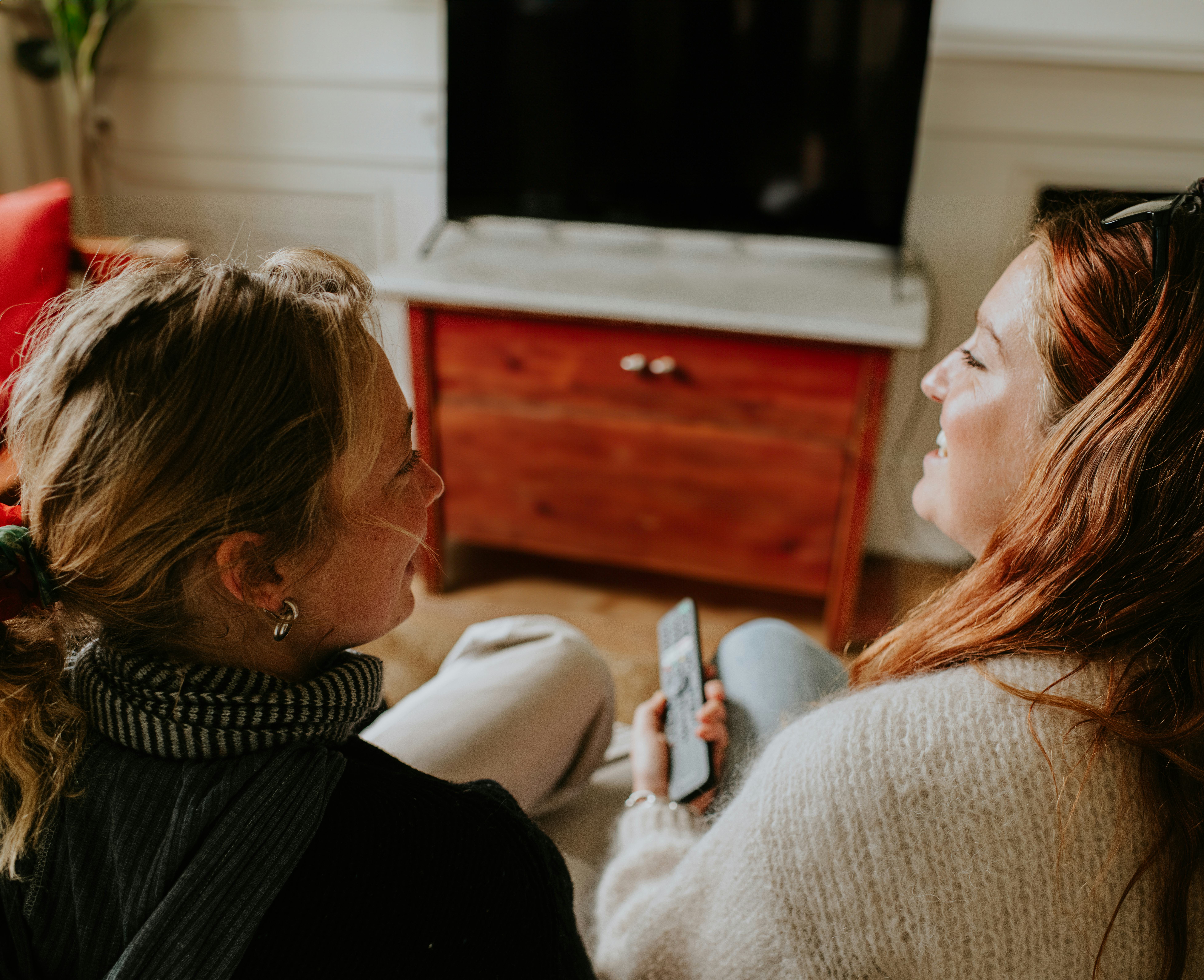 Two woman laugh at silly facts about each other as they watch tv from the couch.