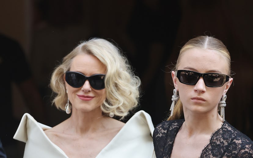 Two women in stylish attire exiting a building, one in a white dress and sunglasses, the other in a ...