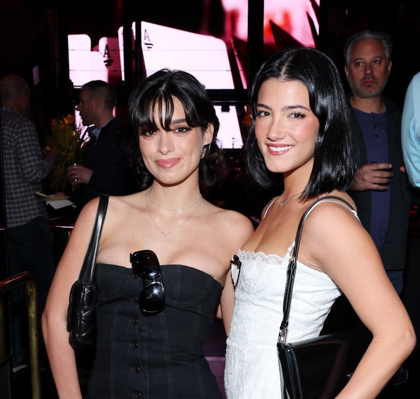 Two women smiling in a club, one in a black dress and the other in white, both wearing small purses.