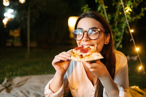 A woman wearing eyeglasses eating a slice of pizza outdoors while watching a movie with her friends...