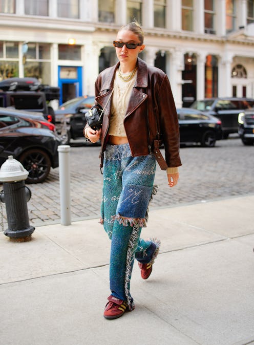Gigi Hadid wears a brick red leather jacket, chunky knit top, fringe pants, and Adidas x Wales Bonne...