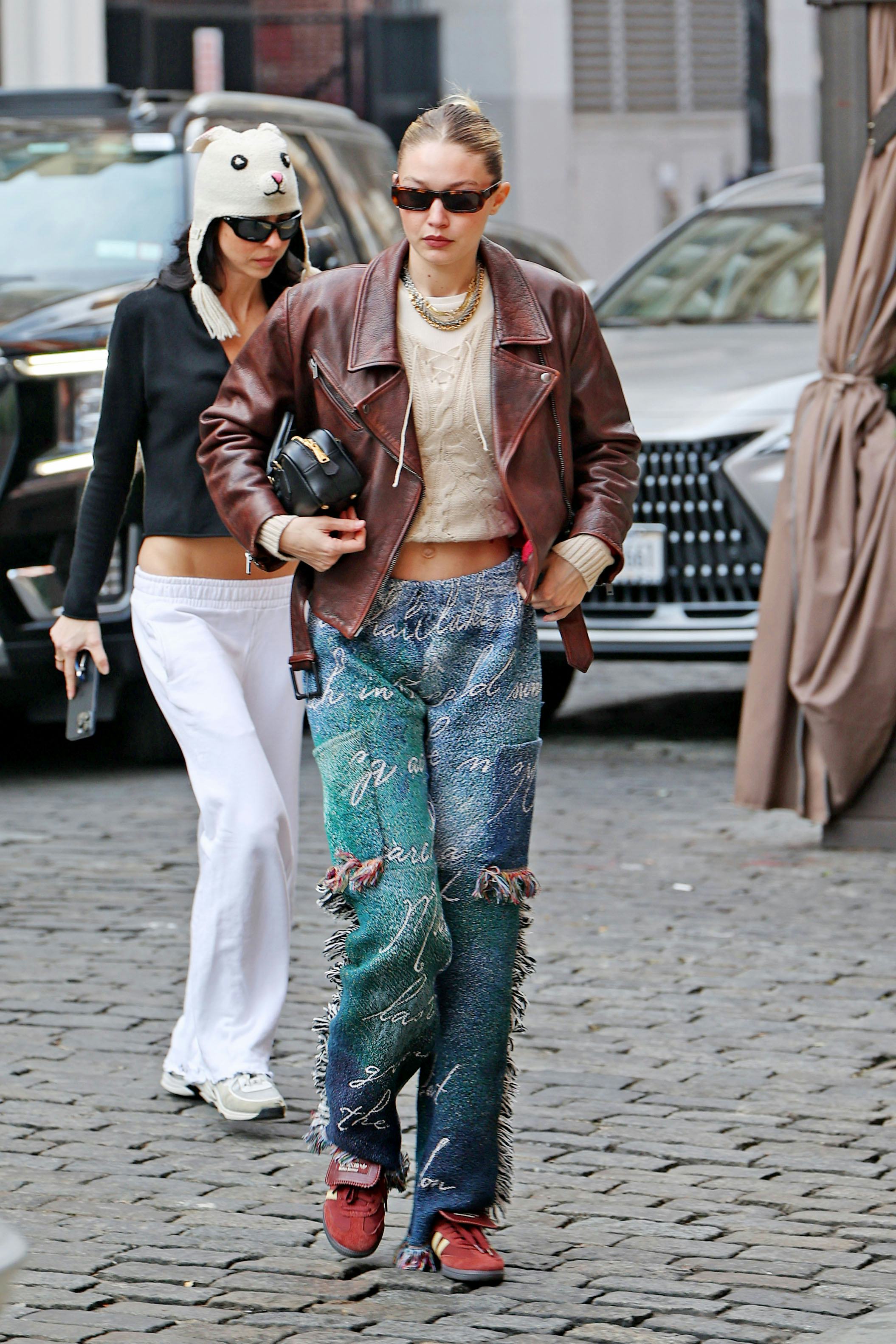 Gigi Hadid wears a brick red leather jacket, chunky knit top, fringe pants, and Adidas x Wales Bonne&hellip;