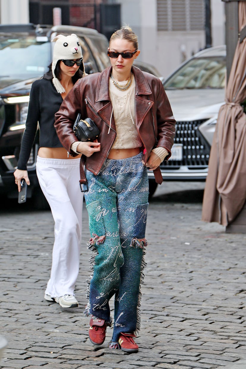 Gigi Hadid wears a brick red leather jacket, chunky knit top, fringe pants, and Adidas x Wales Bonne…