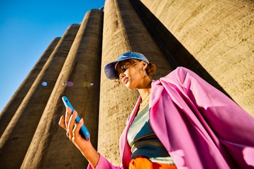 A woman in a bright pink jacket holds a smartphone, standing near tall, columnar structures under a ...