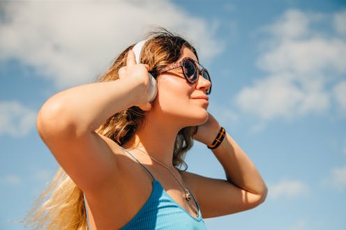 A young woman wearing sunglasses enjoys music on her headphones under a clear blue sky.