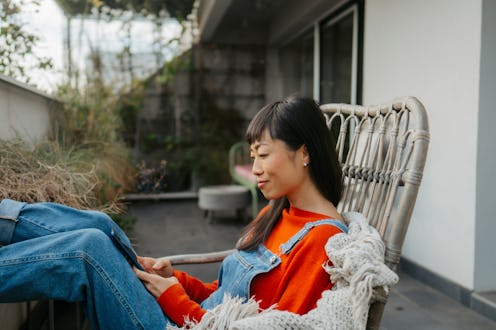 Photo of a smiling Japanese woman using a smartphone while sitting on the balcony of her loft apartm...
