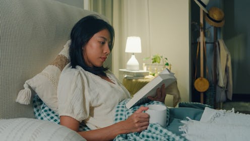 Young Asian girl reading book and drinking milk sitting on the bed in home at night. Insomnia, Cyber...