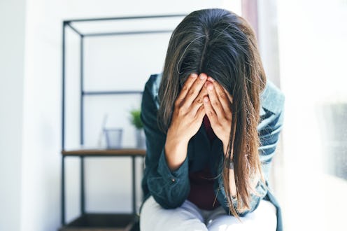 A woman sits with her head in her hands, wearing a teal jacket and a red shirt, in an office with su...