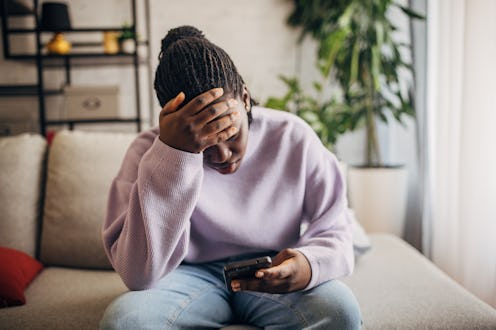Young black woman looks stressed while reading a message on her smartphone in her living room