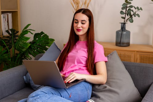 Young woman smiles as she works on her laptop while sitting on a cozy sofa