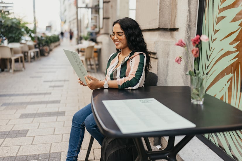 A young woman sitting at a table outside a cafe reading the menu and looking at jokes for adults