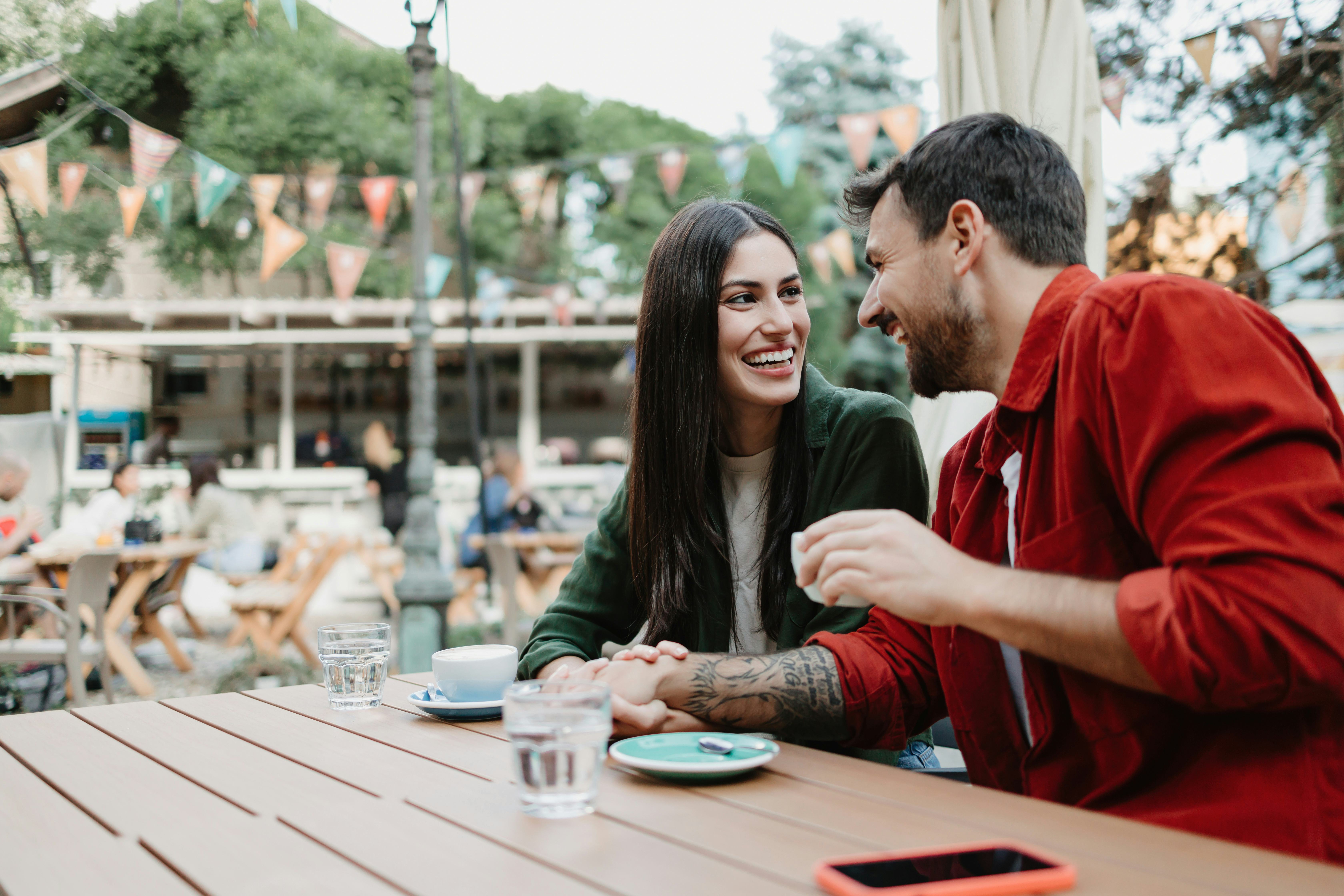 A young couple sit at an outdoor table having coffee laughing at fun pickup lines