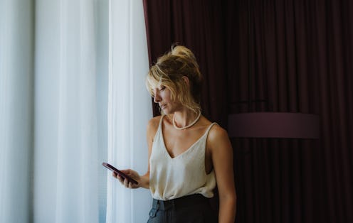 Portrait of an elegant serious woman standing by the window of her hotel room, looking down at her m...