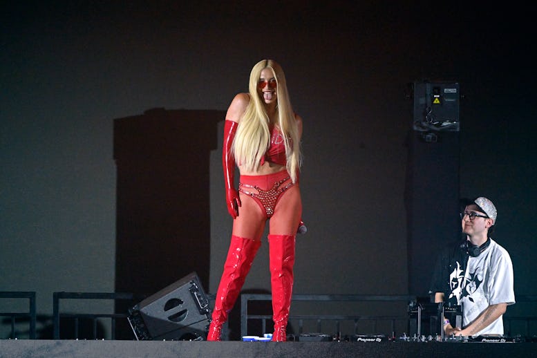 A performer in a red outfit with long blonde hair and red boots on stage, with a male DJ in the back...