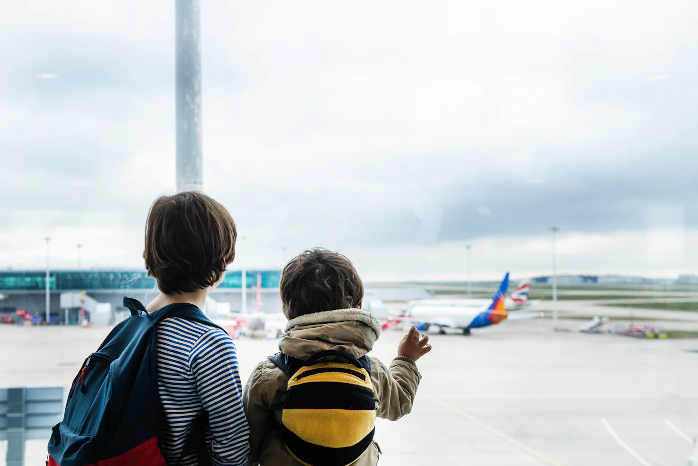 Two people, one adult and one child, look out a large airport window at planes on tarmac, child pointing outside.