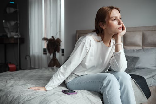 Phone anxiety. Upset woman sitting on bed with phone looking out window, feeling depressed, waiting ...