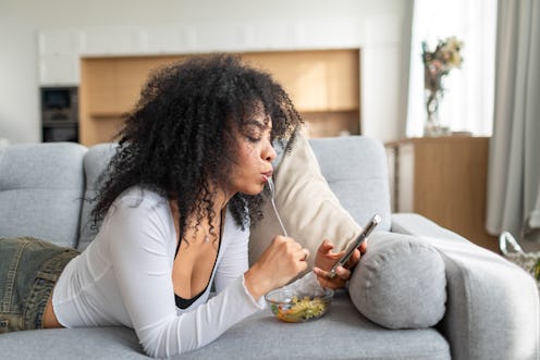 Young multiracial woman lying down on sofa with mobile and eating pasta salad at home