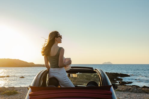 Cheerful young woman in an open-top car. She is enjoying the sunset and looking at the sea