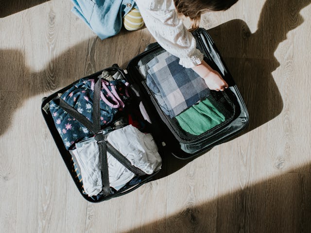 Top down conceptual image of a child kneeling on the floor. She opens a neatly packed suitcase, fill...