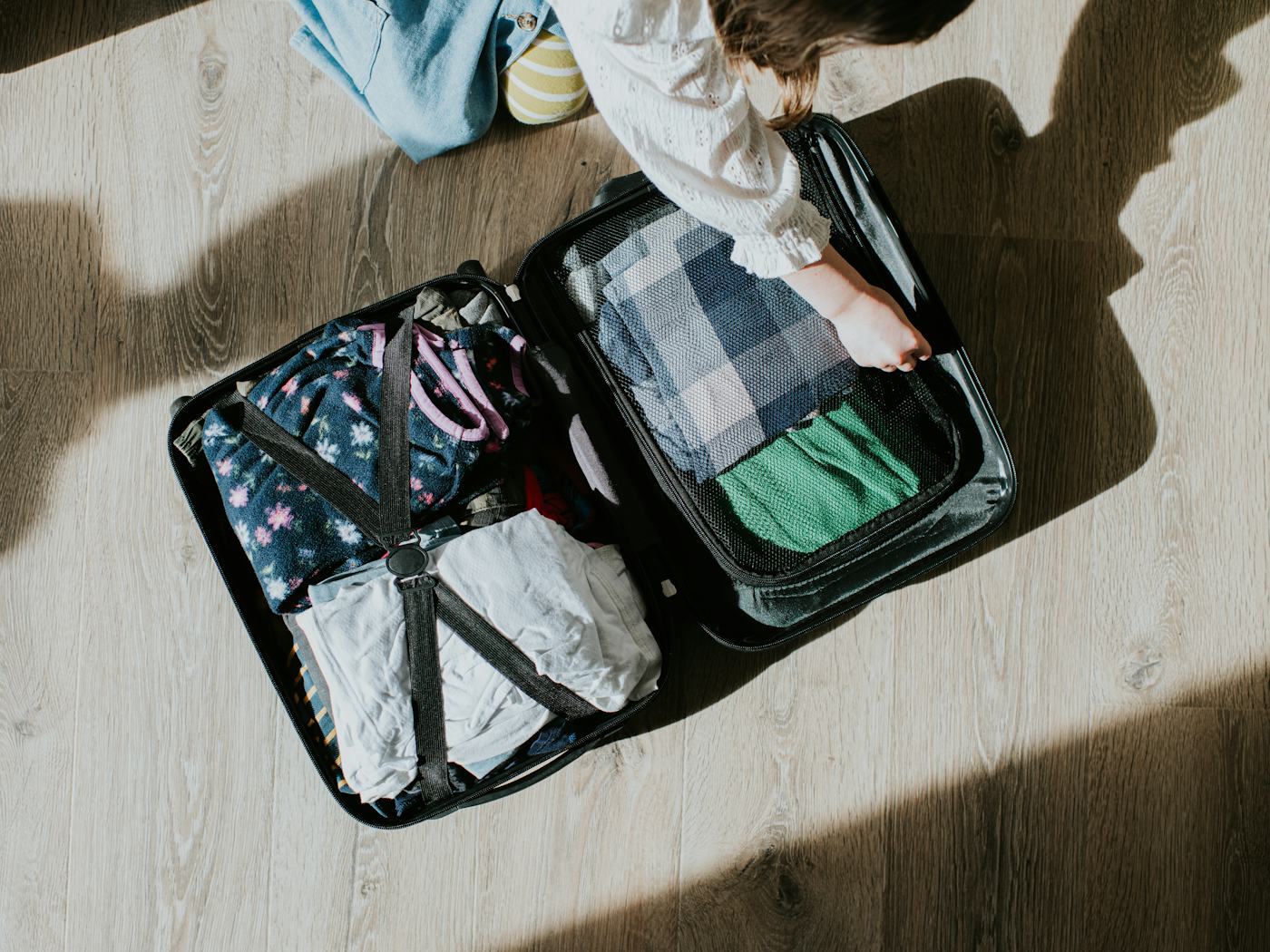 Top down conceptual image of a child kneeling on the floor. She opens a neatly packed suitcase, filled with clean, folded clothing.