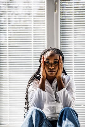 Portrait of overworked female doctor sitting on the floor, by the window, head in hands, looking at ...